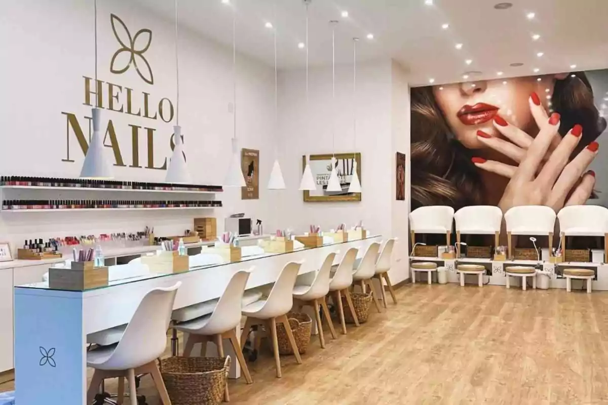 Un salón de manicura moderno con sillas blancas, mesas de trabajo organizadas y una gran imagen de una mujer con uñas rojas en la pared.