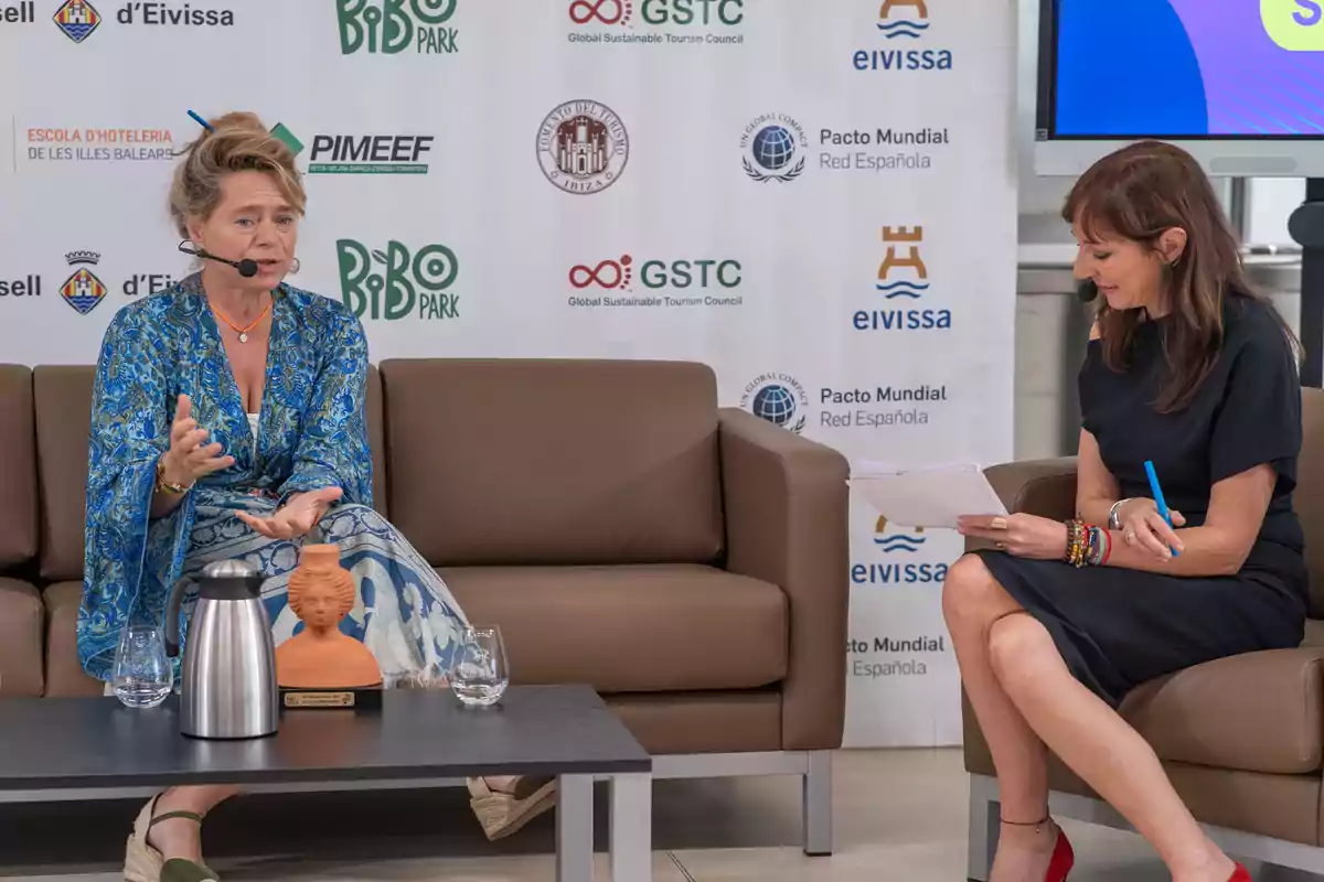 Dos mujeres conversan en un evento sentadas en un sofá con un fondo de logotipos de patrocinadores. Una de ellas lleva un vestido azul y habla mientras la otra toma notas. En la mesa frente a ellas hay una jarra de metal, una pequeña escultura y vasos de agua.