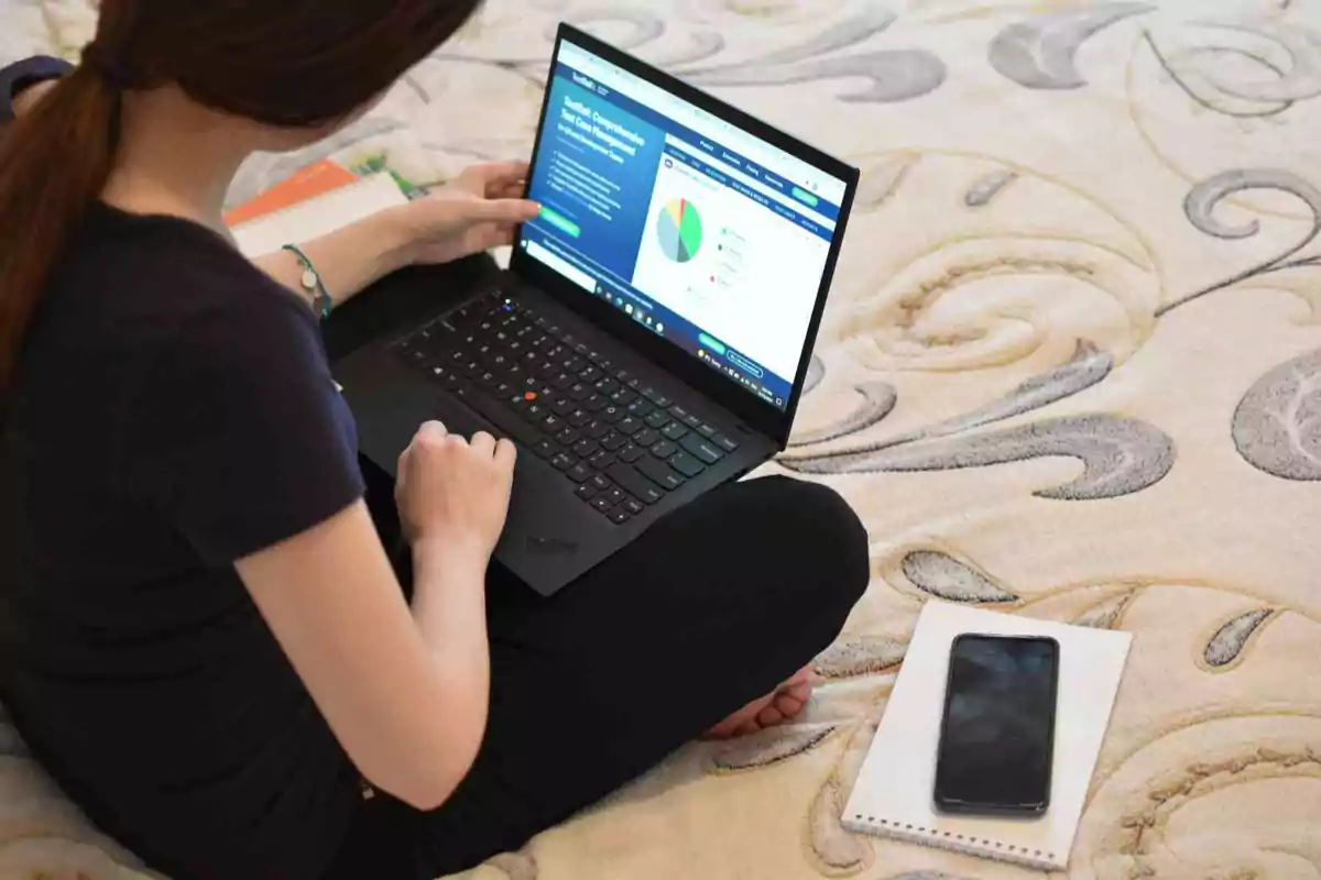Una persona sentada en el suelo usando una laptop con un teléfono móvil y un cuaderno al lado.