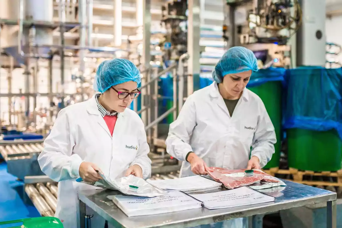 Dos trabajadoras con batas blancas y gorros de malla azul revisan documentos y paquetes en una planta industrial.