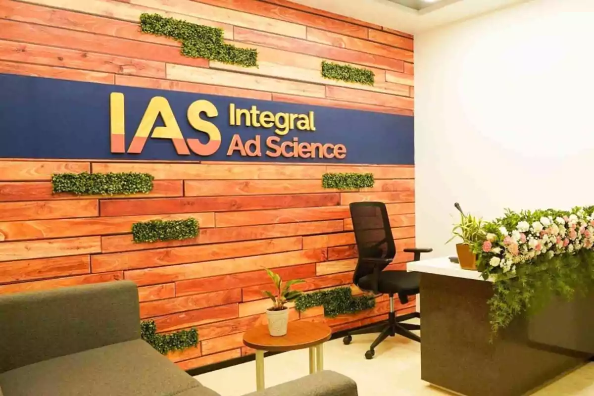 Oficina moderna con un letrero de "IAS Integral Ad Science" en una pared de madera decorada con plantas y un escritorio con flores.