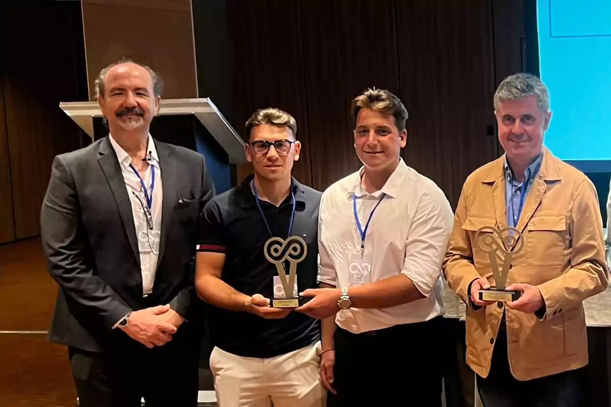 Cuatro hombres posan en un evento, dos de ellos sostienen trofeos y todos llevan gafetes de identificación.