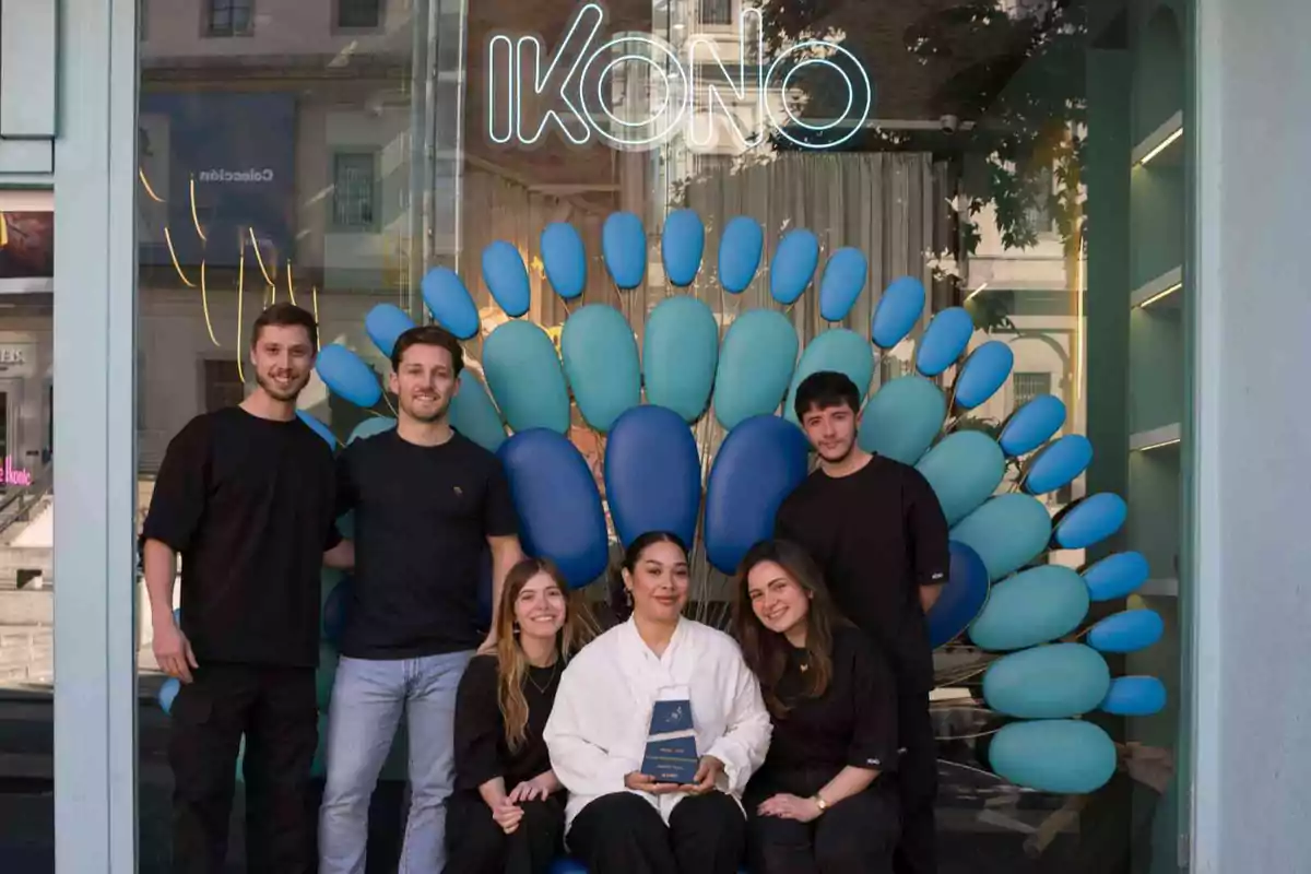 Seis personas posan sonrientes frente a una estructura decorativa azul en forma de abanico bajo un letrero que dice IKONO, una de ellas sostiene un trofeo.
