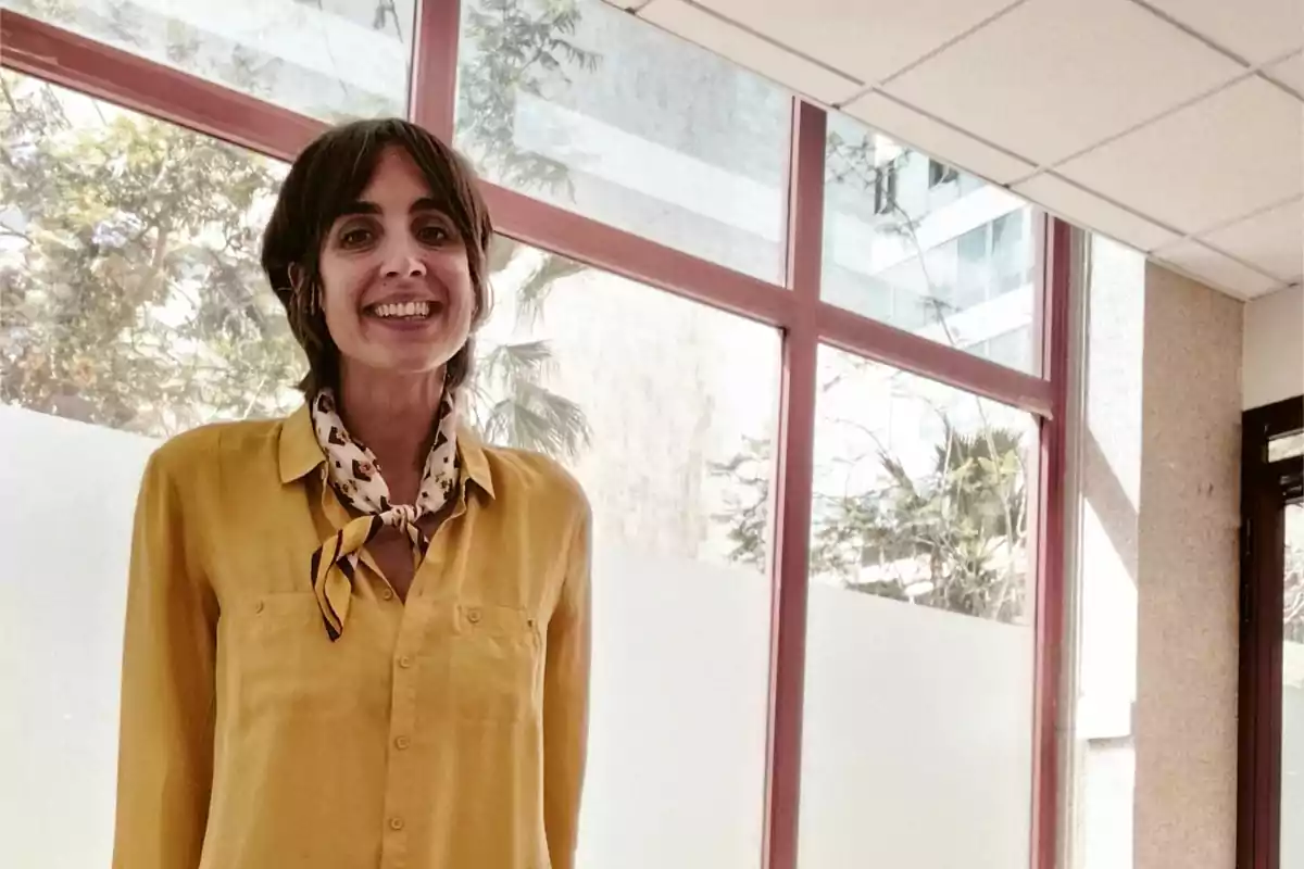 Una persona sonriente con camisa amarilla y pañuelo en el cuello está de pie frente a una ventana grande en un espacio interior iluminado.