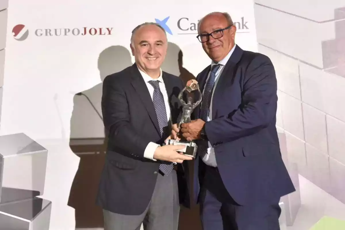 Dos hombres en trajes formales sosteniendo un trofeo en un evento con los logotipos de Grupo Joly y CaixaBank en el fondo.