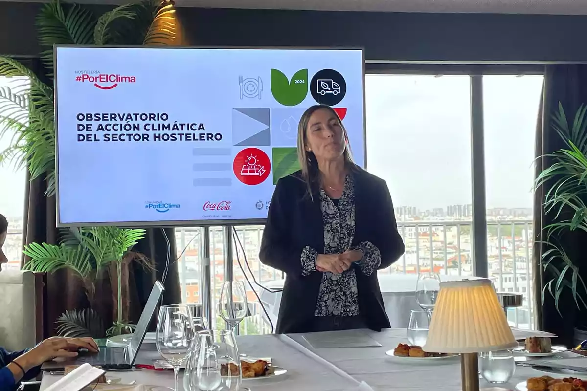 Una mujer de pie frente a una pantalla que muestra información sobre el Observatorio de Acción Climática del Sector Hostelero, con una mesa servida en primer plano.