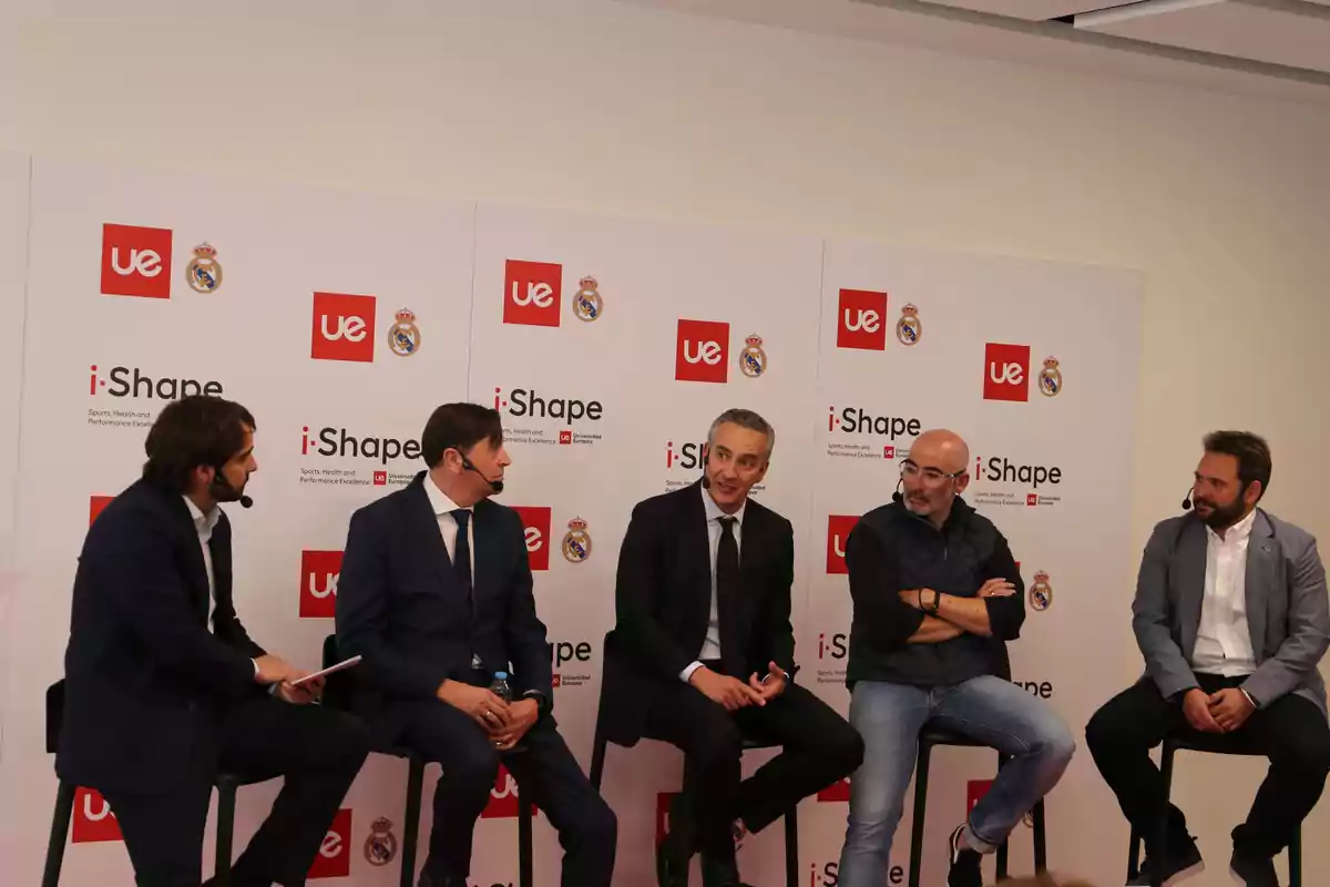 Cinco hombres sentados en sillas participando en una mesa redonda con micrófonos, frente a un fondo con logotipos de i-Shape, UE y el escudo del Real Madrid