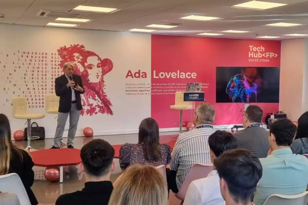 Un hombre está dando una presentación en un evento de tecnología en un auditorio, con una imagen de Ada Lovelace y texto en la pared de fondo, y varias personas sentadas escuchando.