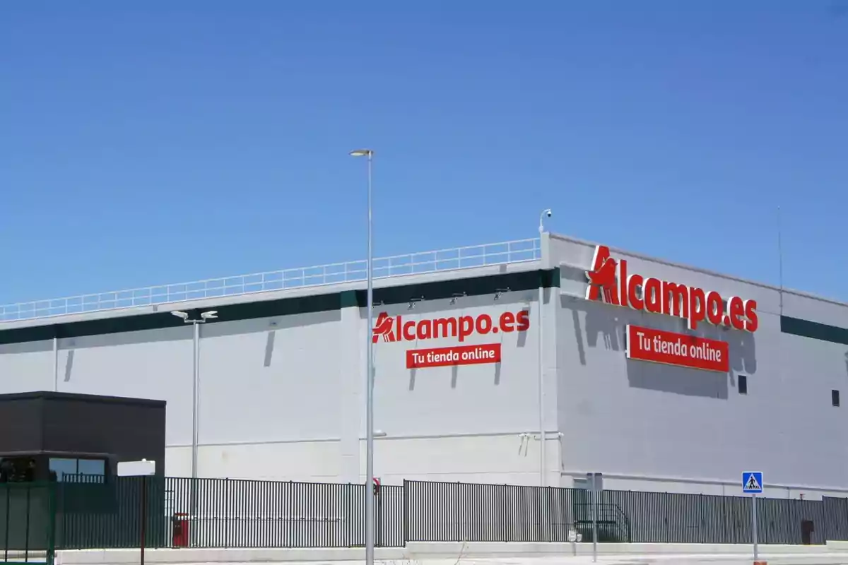 Fachada de un almacén de Alcampo.es con letreros que dicen "Tu tienda online" en un día soleado.