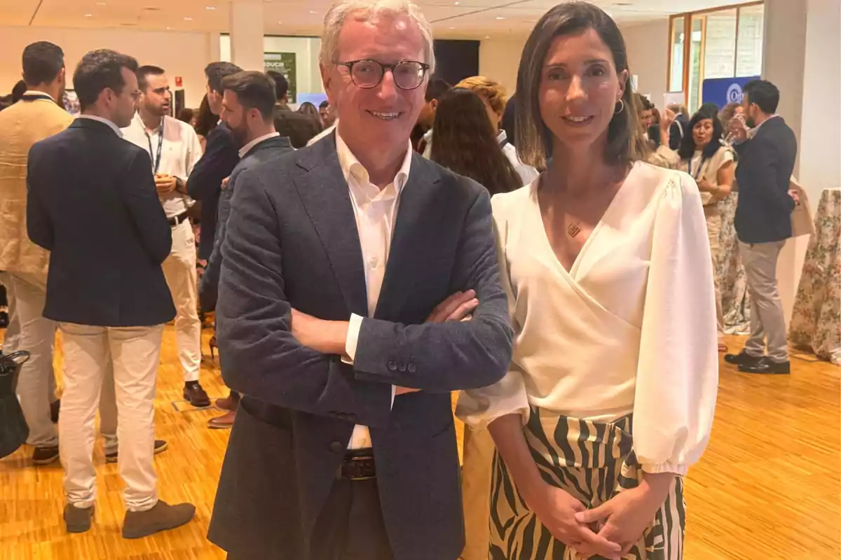 Dos personas posan sonrientes en primer plano en un evento social o profesional, rodeados de otras personas conversando en un espacio interior iluminado.