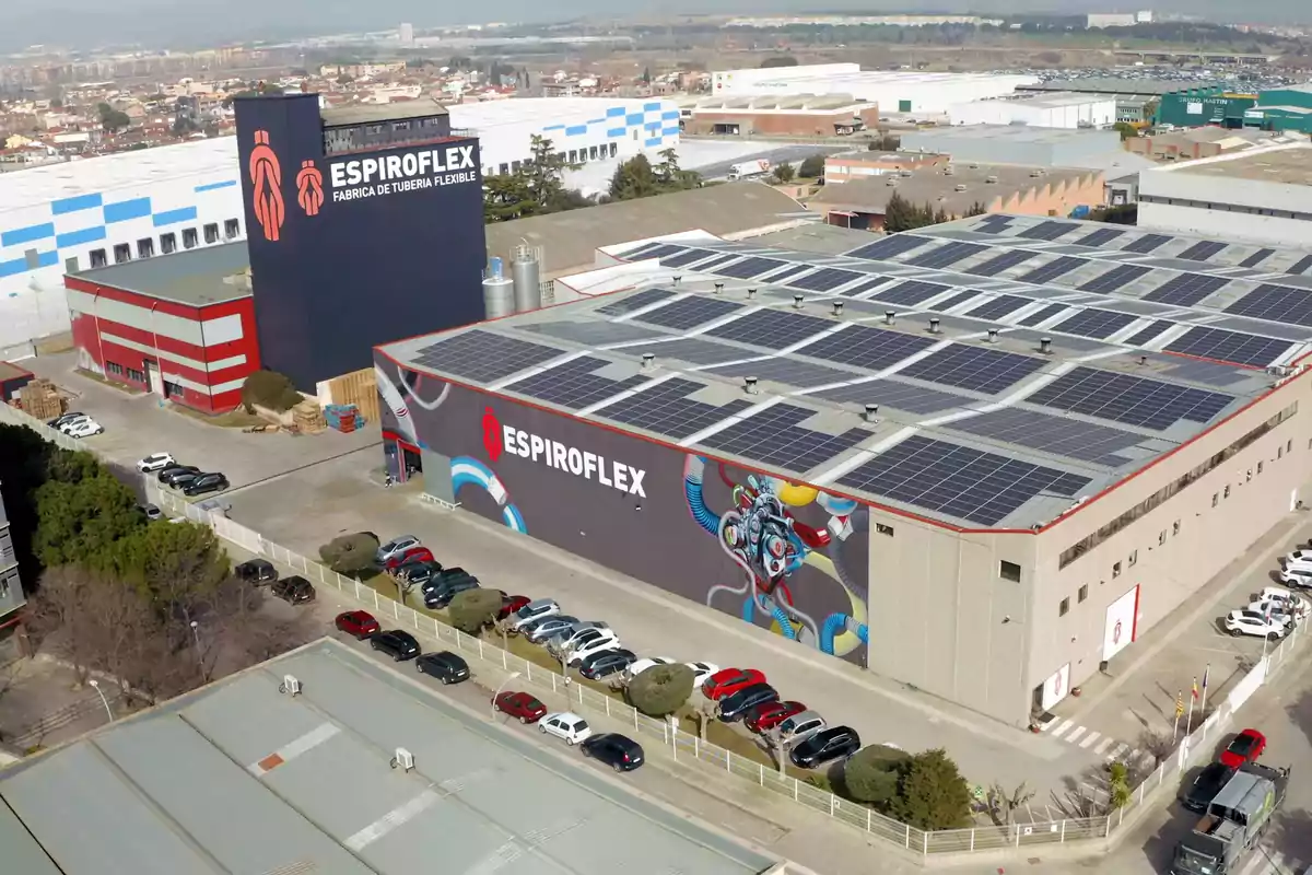 Vista aérea de una fábrica con paneles solares en el techo y un mural colorido en la fachada, rodeada de coches estacionados y otras instalaciones industriales.