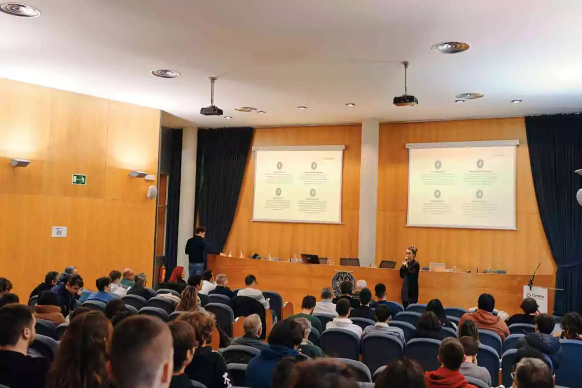 Una conferencia en un auditorio con personas sentadas mientras un presentador habla frente a dos pantallas con diapositivas.