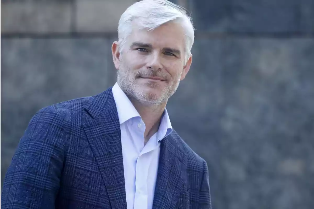 Hombre de cabello canoso y barba incipiente, vestido con camisa blanca y saco azul, sonriendo frente a un fondo borroso.