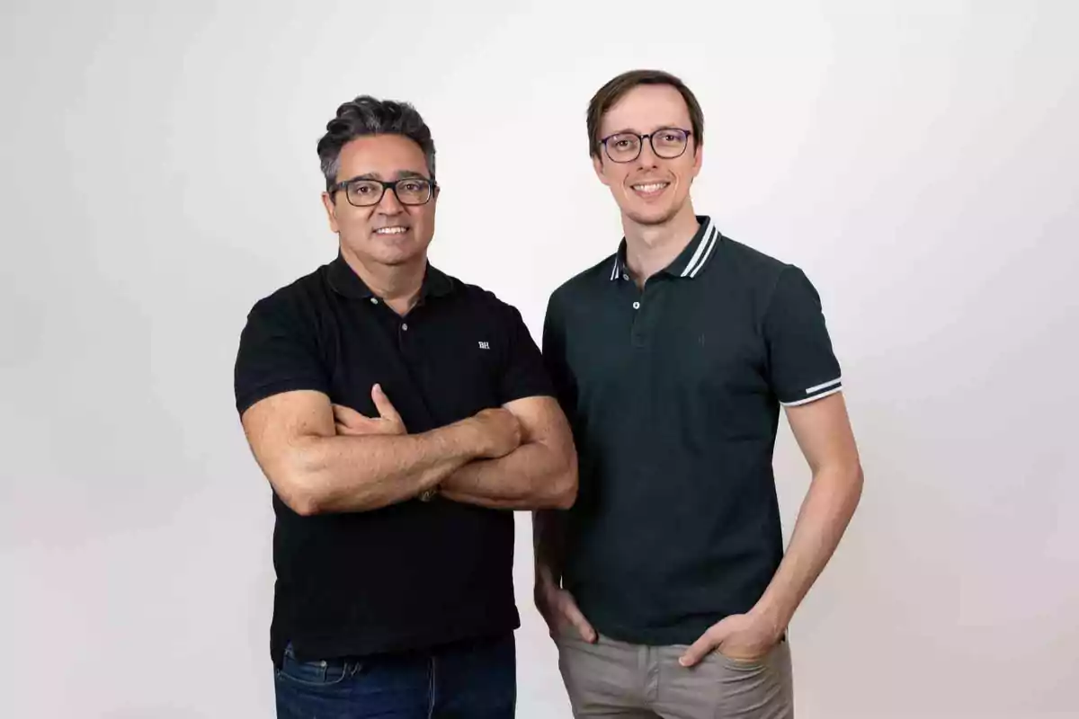 Dos personas de pie sonriendo con camisas de polo y gafas en un fondo blanco.