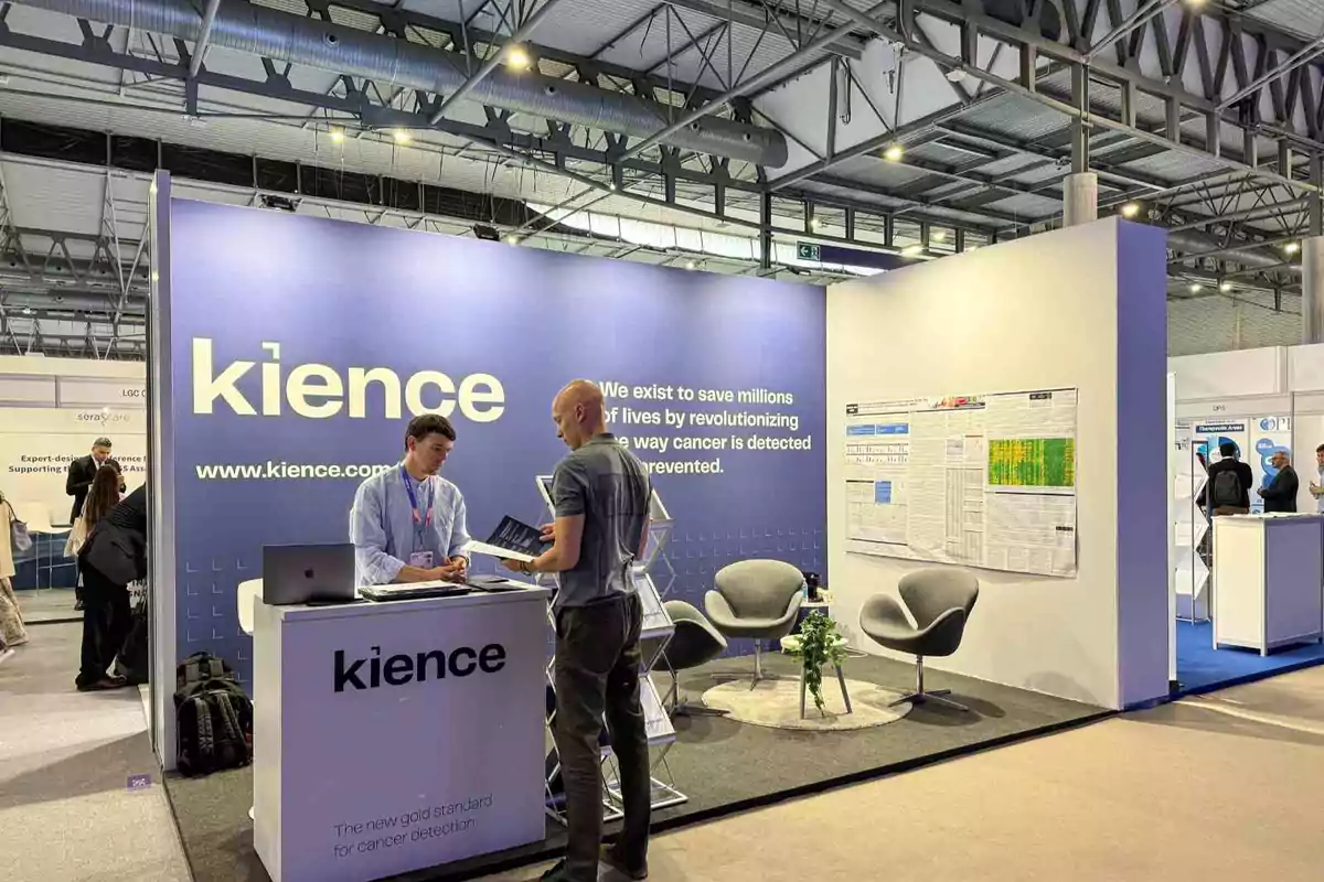 Un stand de exhibición con el nombre "Kience" en un evento, donde dos personas están conversando frente a un mostrador, con sillas y una mesa en el área de descanso, y un cartel informativo en la pared.