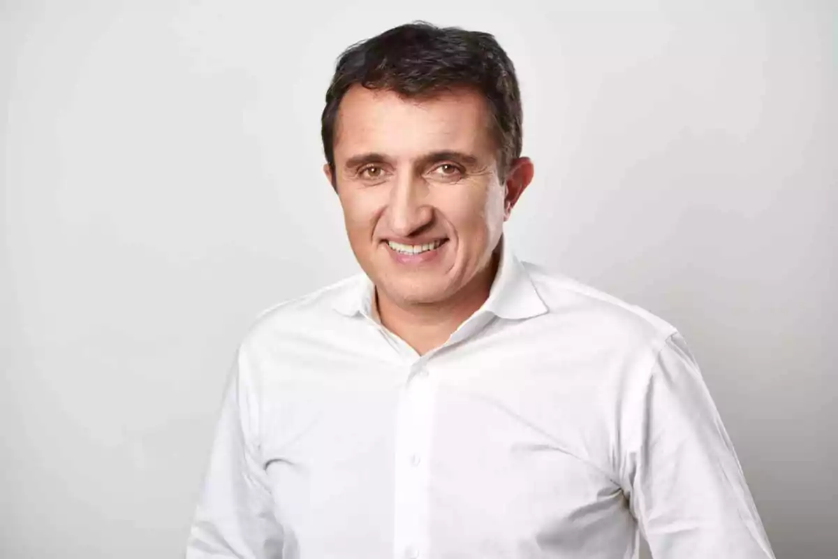 Hombre sonriendo con camisa blanca sobre fondo gris.