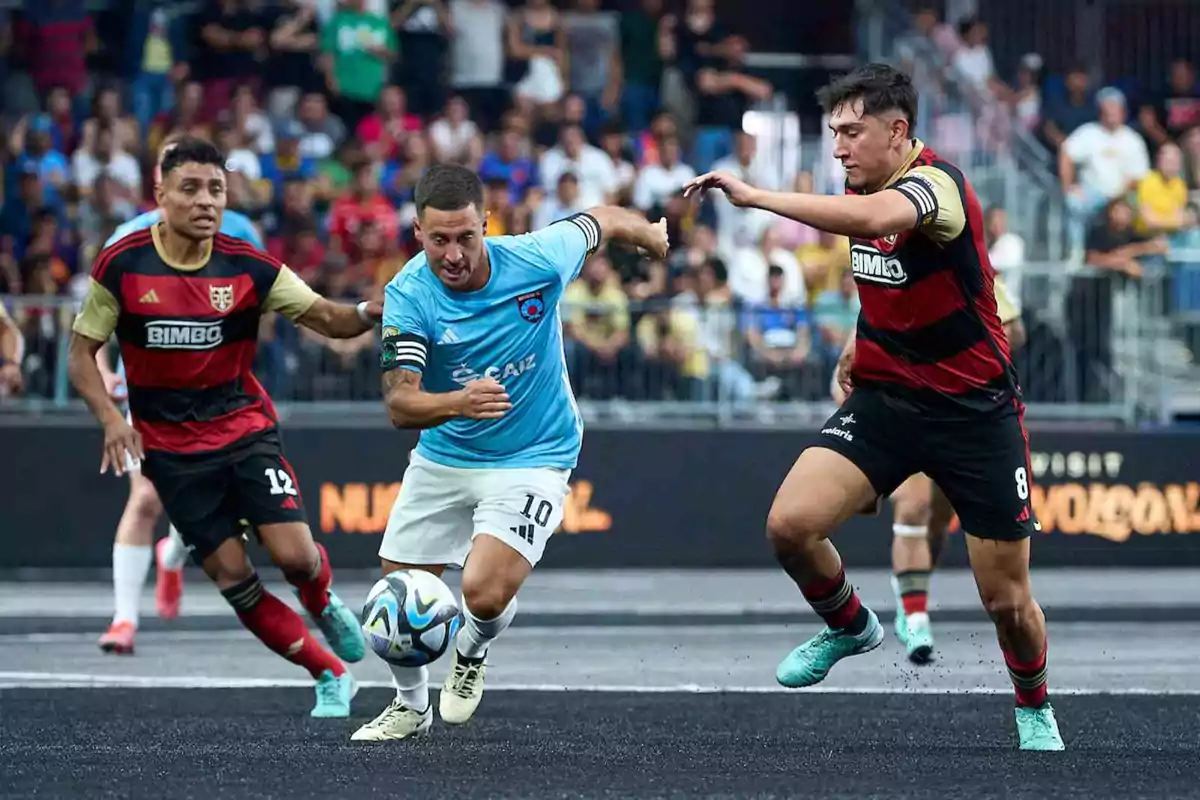 Jugadores de fútbol compiten por el balón en un partido, uno con uniforme azul y los otros con uniforme rojo y negro, mientras el público observa desde las gradas.