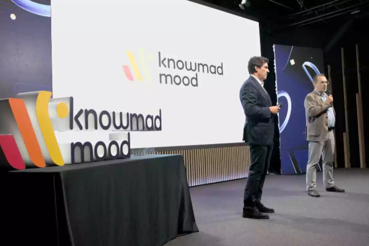 Dos personas en un escenario presentan frente a una pantalla grande que muestra el logo de "knowmad mood", con un letrero del mismo logo sobre una mesa a la izquierda.
