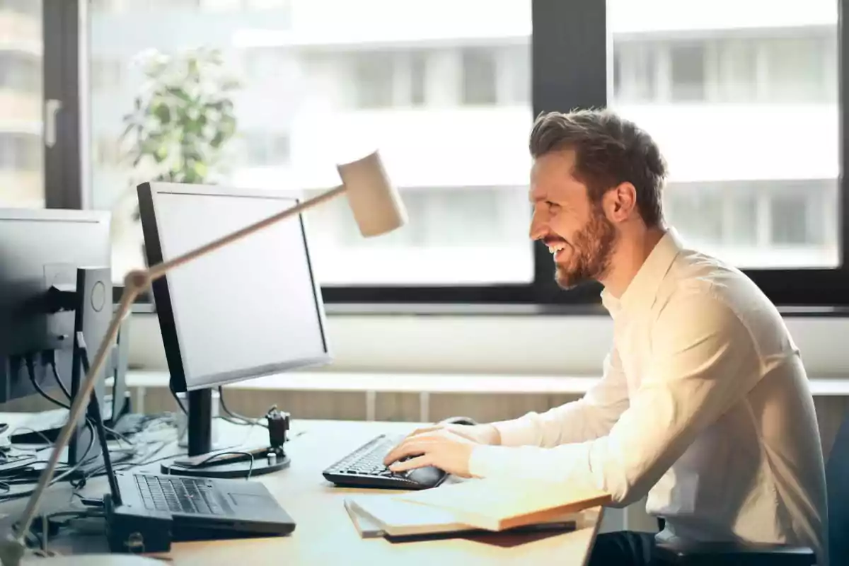 Hombre sonriendo mientras trabaja en una computadora en una oficina iluminada por la luz natural.