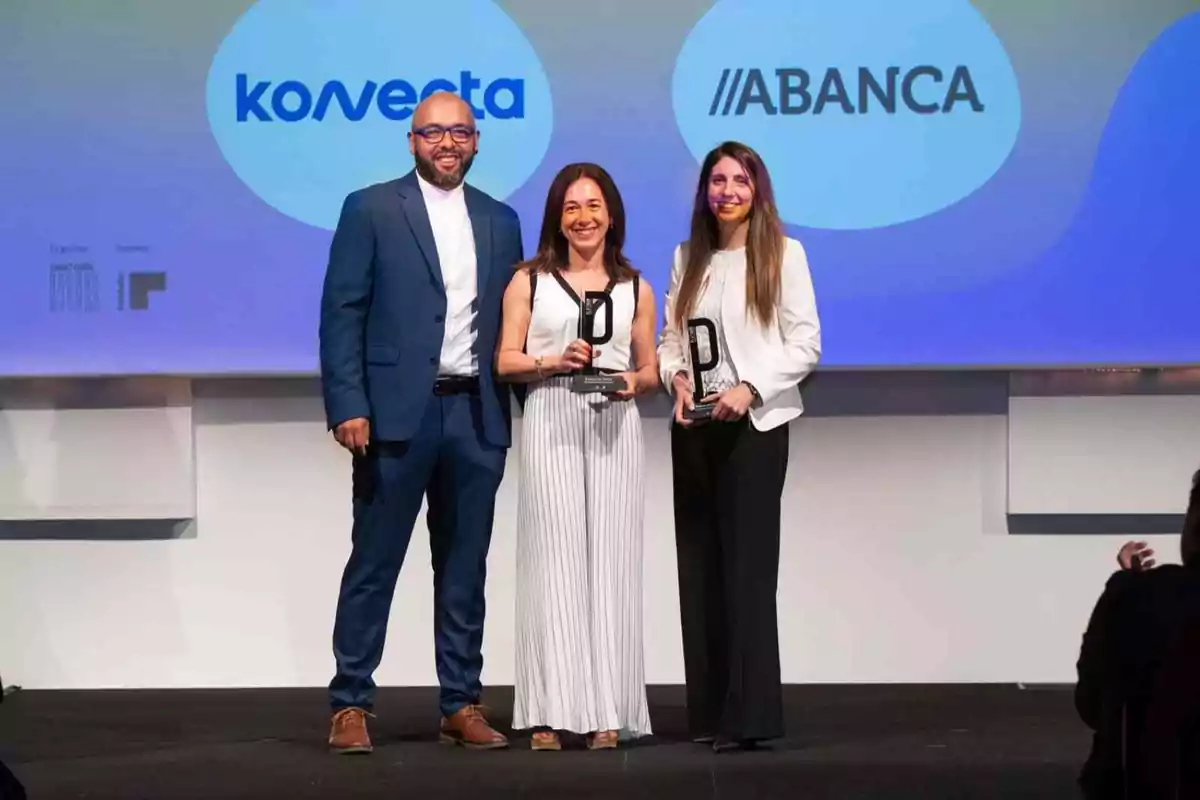 Tres personas posan sonrientes en un escenario durante una ceremonia de premiación, dos de ellas sostienen trofeos y al fondo se ven los logotipos de Konecta y Abanca.