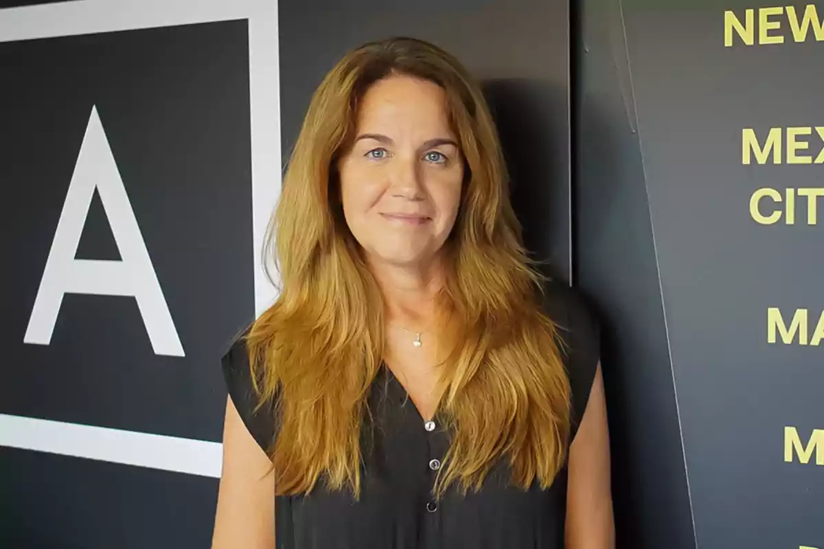 Mujer de cabello largo y rubio posando frente a una pared con letras y un símbolo de "A".