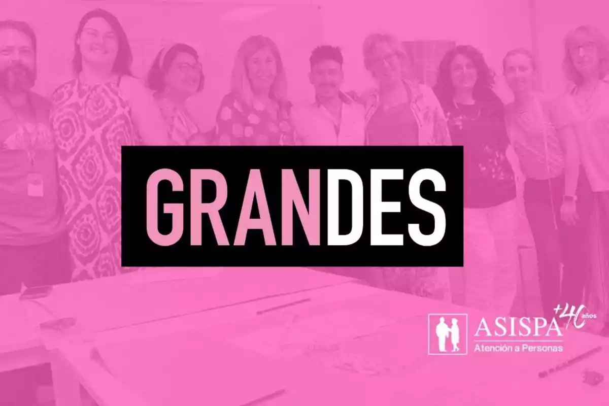 Un grupo de personas sonríe frente a una mesa con la palabra "GRANDES" destacada en el centro sobre un fondo rosa.