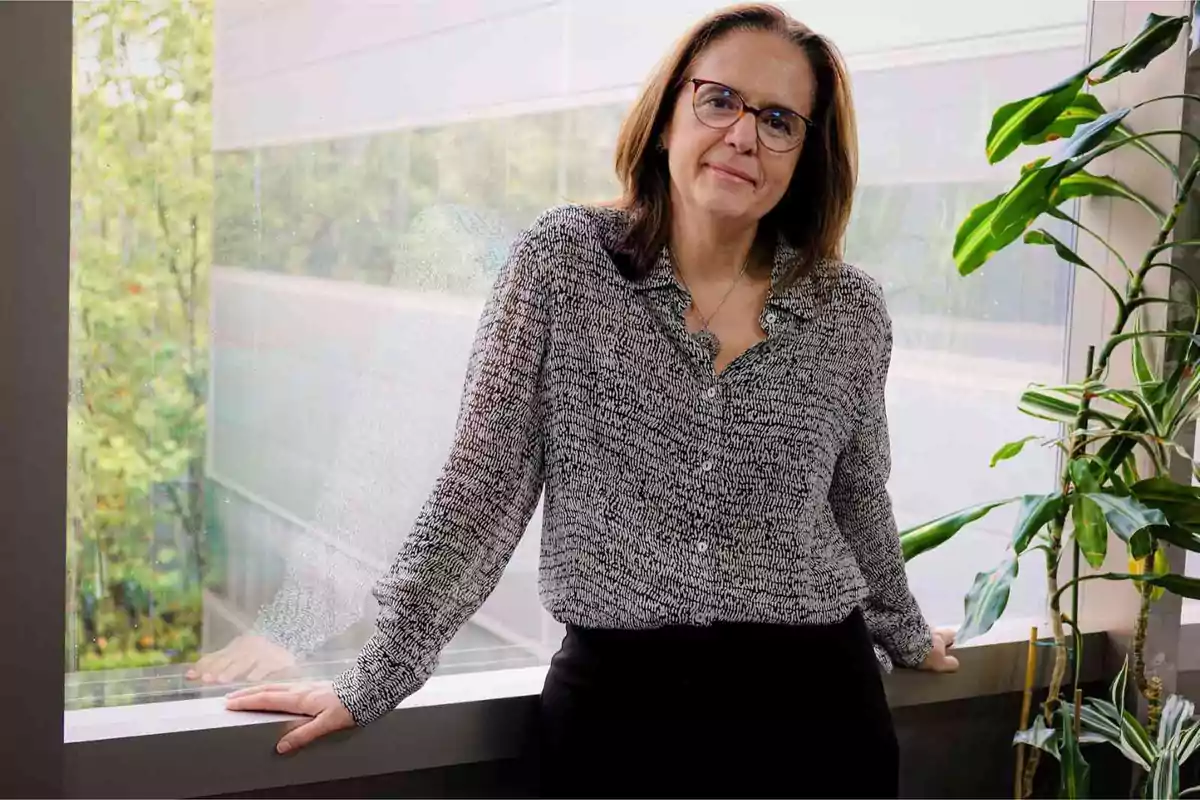 Una mujer con gafas y camisa estampada está de pie junto a una ventana con plantas al lado.