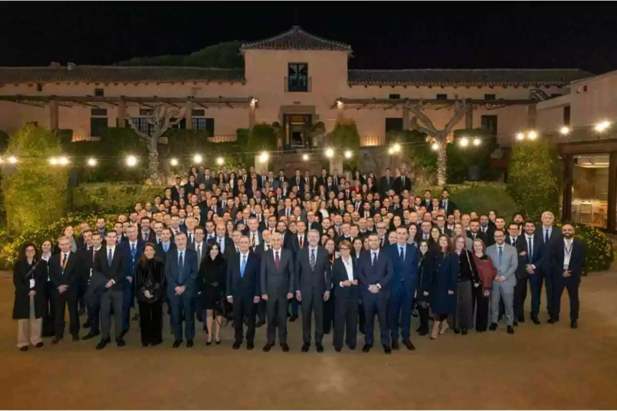 Un gran grupo de personas posando en un evento formal al aire libre frente a un edificio iluminado por la noche.