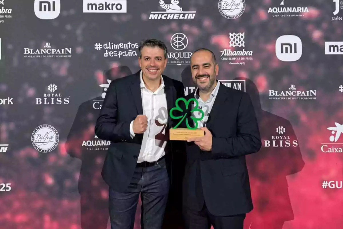 Dos hombres posan en un evento con un fondo lleno de logotipos de marcas uno de ellos sostiene un trofeo en forma de trébol verde.