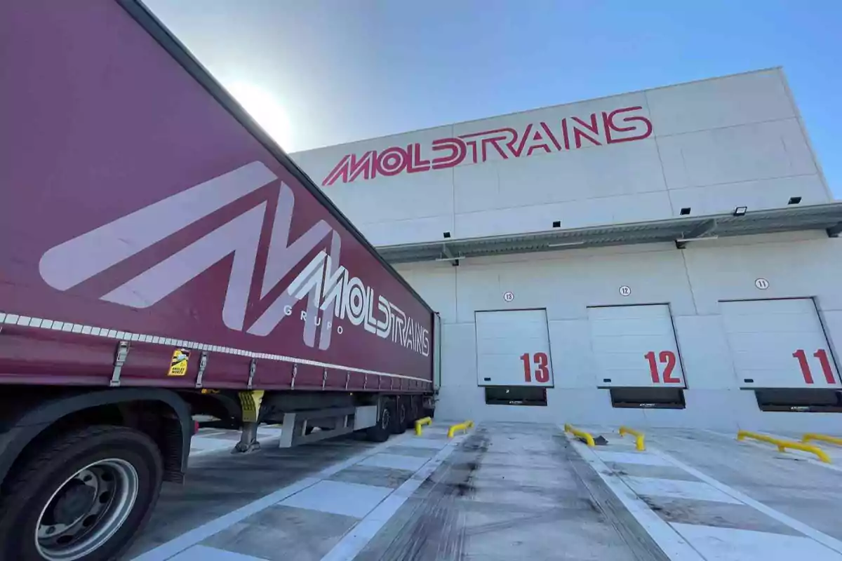 Camión de carga estacionado frente a un edificio con el logo de Moldtrains y varias puertas de carga numeradas.