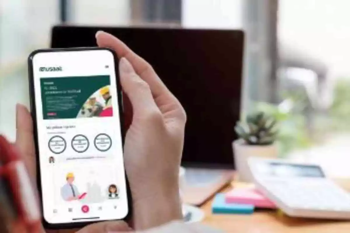 Una persona sostiene un teléfono móvil que muestra una aplicación mientras está sentada en un escritorio con una computadora portátil y una calculadora.