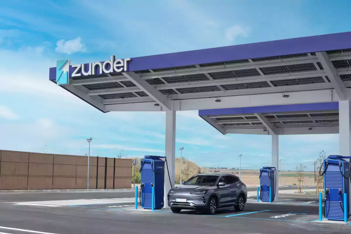 Estación de carga para vehículos eléctricos con un coche gris estacionado bajo un techo con paneles solares y cargadores azules.