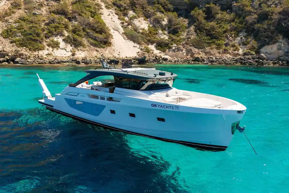 Un yate blanco de OK YACHTS anclado en aguas cristalinas cerca de una costa rocosa con vegetación.