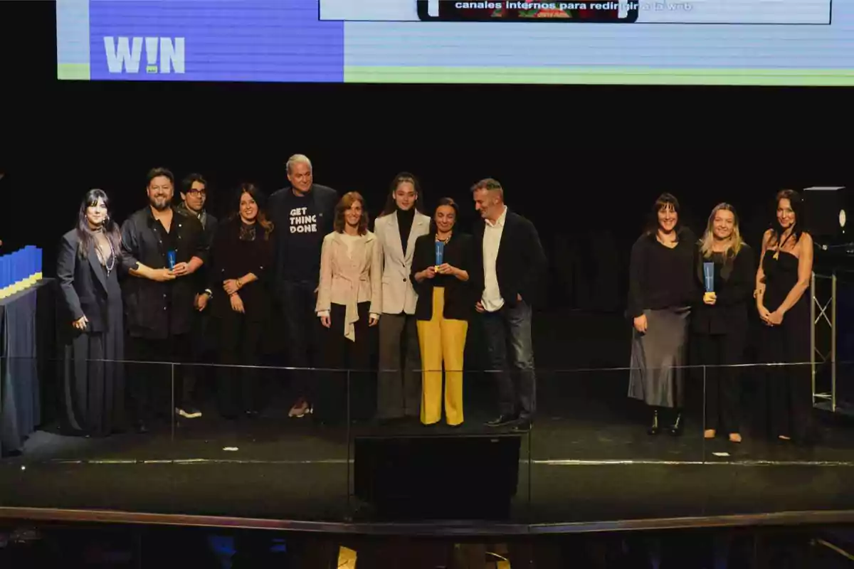 Un grupo de personas posando en un escenario durante un evento, algunas sosteniendo premios, con una pantalla de fondo que muestra la palabra "WIN".