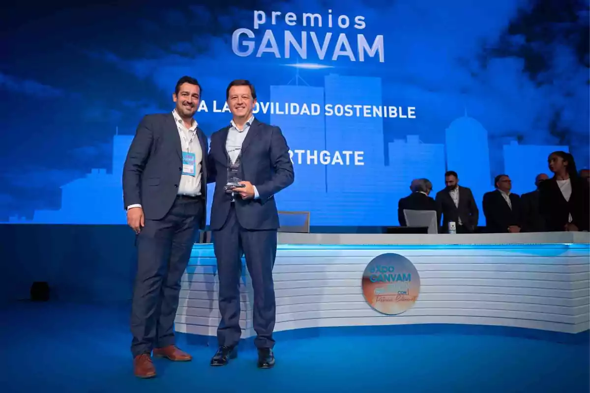 Dos personas posan en un escenario durante los premios GANVAM a la movilidad sostenible, con un fondo azul y un grupo de personas al fondo.