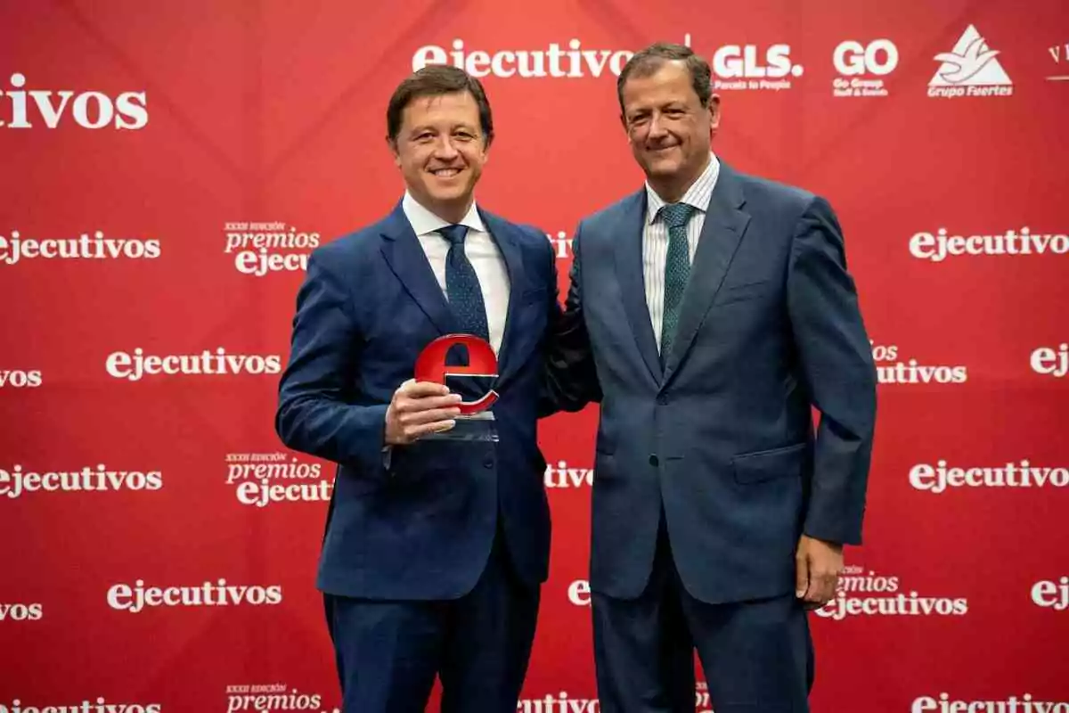 Dos hombres en trajes formales posan frente a un fondo rojo con logotipos de "ejecutivos", uno de ellos sostiene un trofeo.