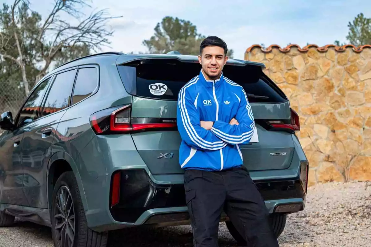 Un hombre con chaqueta azul está apoyado en la parte trasera de un SUV verde con un fondo de pared de piedra.