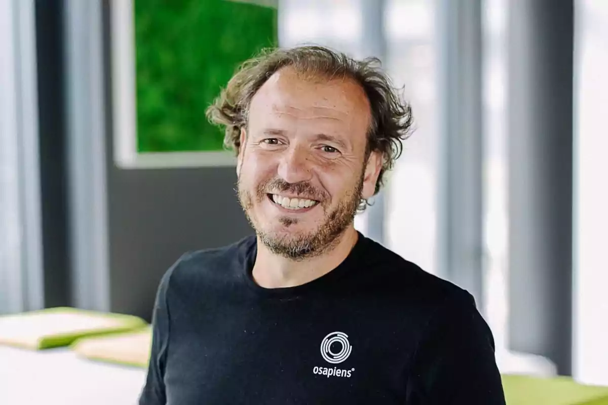 Hombre sonriendo con camiseta negra de osapiens en un entorno interior con fondo desenfocado.