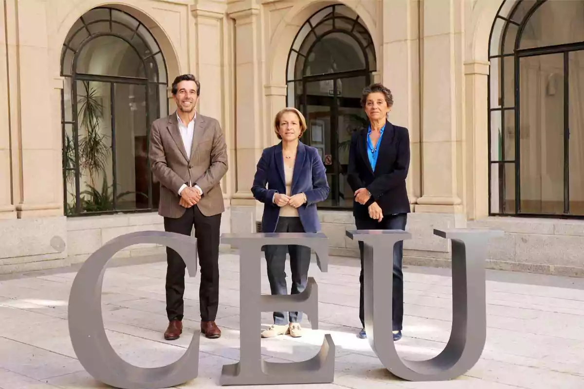 Tres personas posan detrás de grandes letras que forman la palabra CEU en un patio con arcos y ventanas.