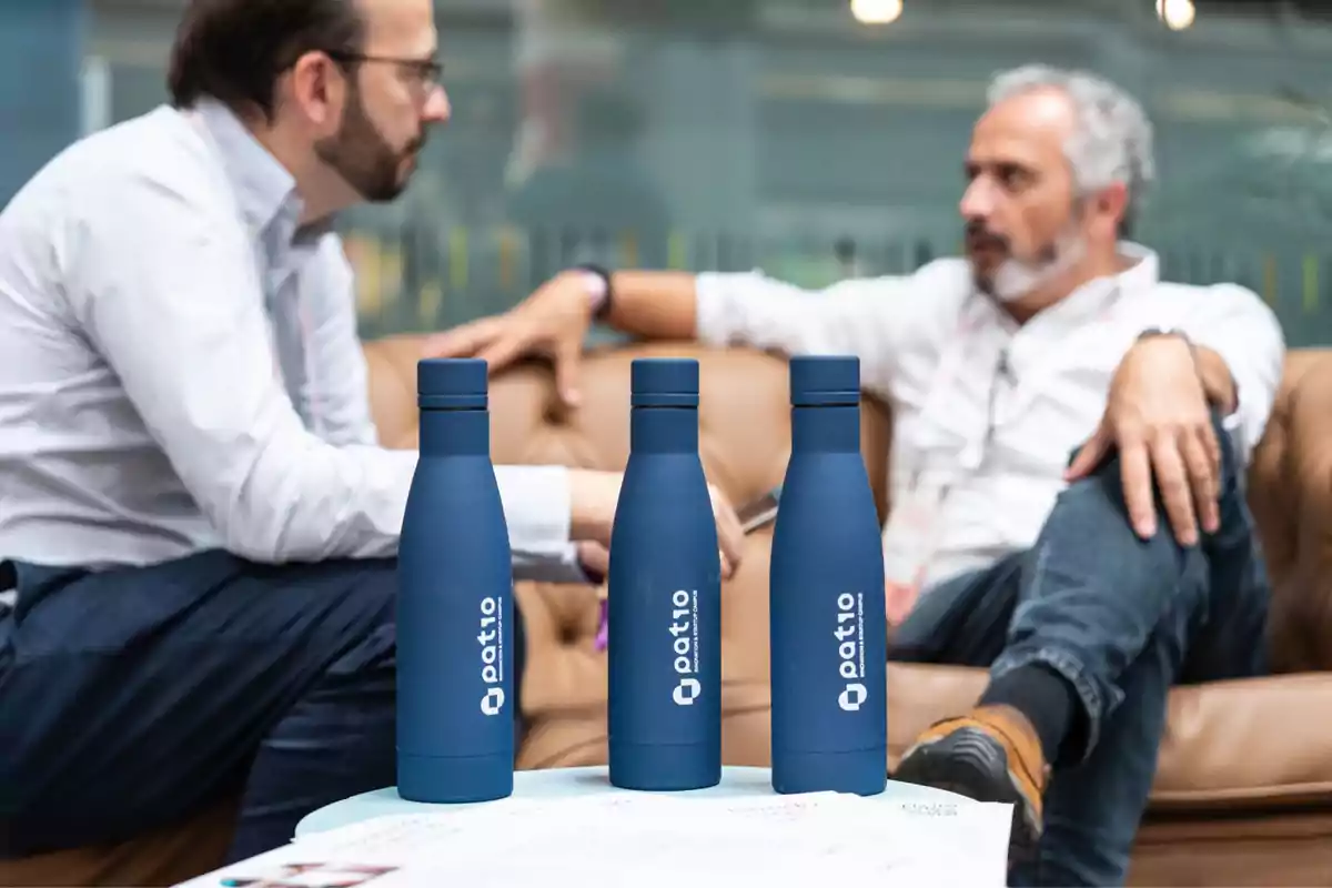 Dos hombres conversando en un sofá con tres botellas azules de la marca "Patio" en primer plano.