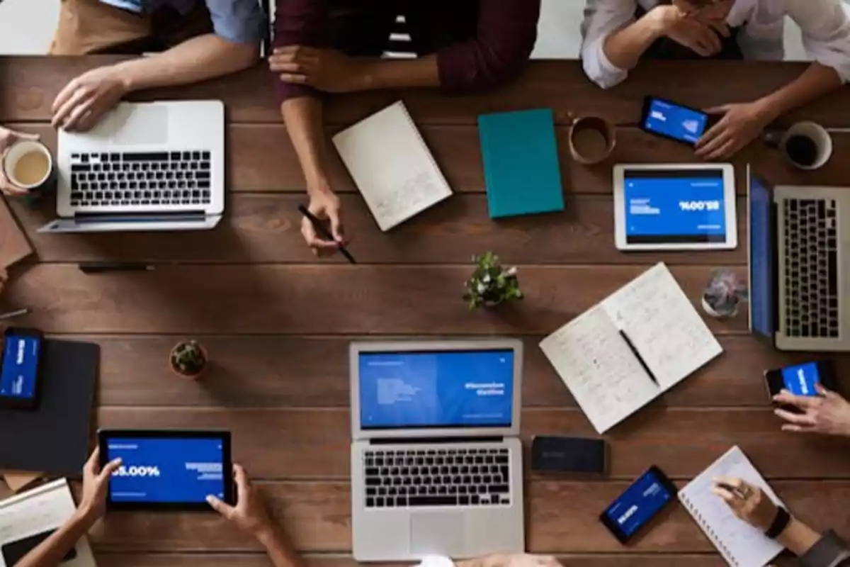 Personas trabajando en una mesa con laptops, tabletas, teléfonos y cuadernos.