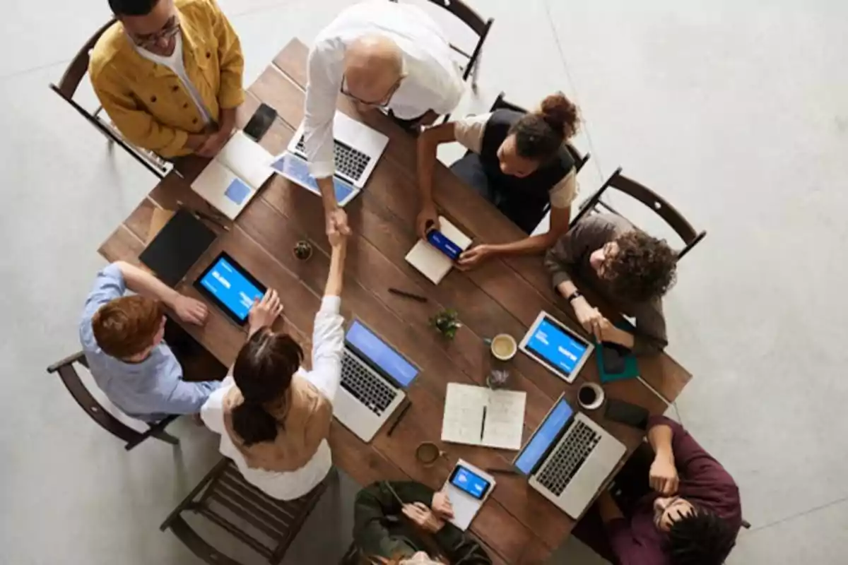 Personas reunidas alrededor de una mesa de madera con laptops y tabletas, dos personas se dan la mano.