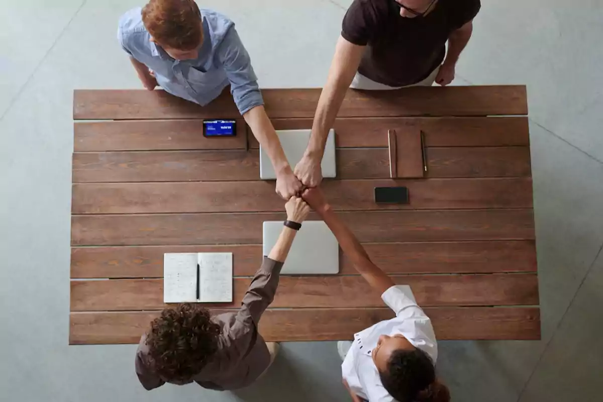 Vista superior de cuatro personas uniendo sus puños sobre una mesa de madera en un gesto de trabajo en equipo.