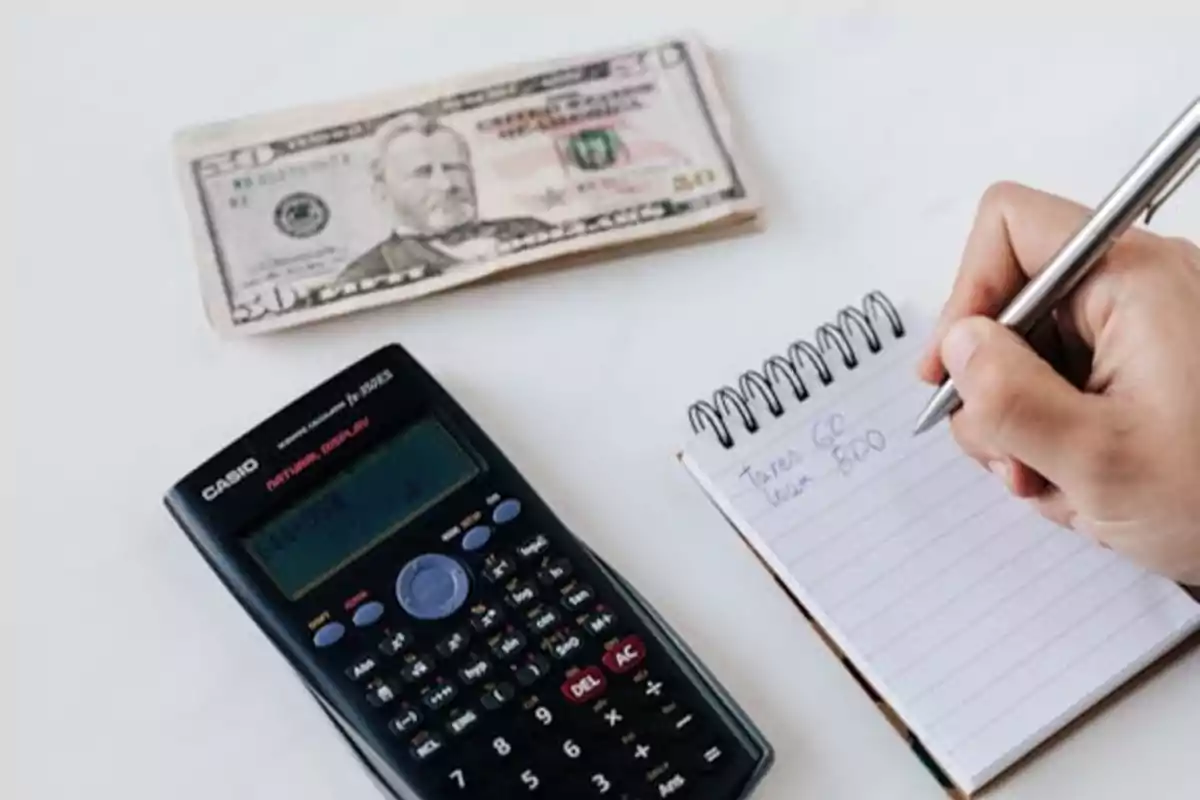 Una persona escribiendo en una libreta con una calculadora y un billete de 50 dólares sobre una mesa.