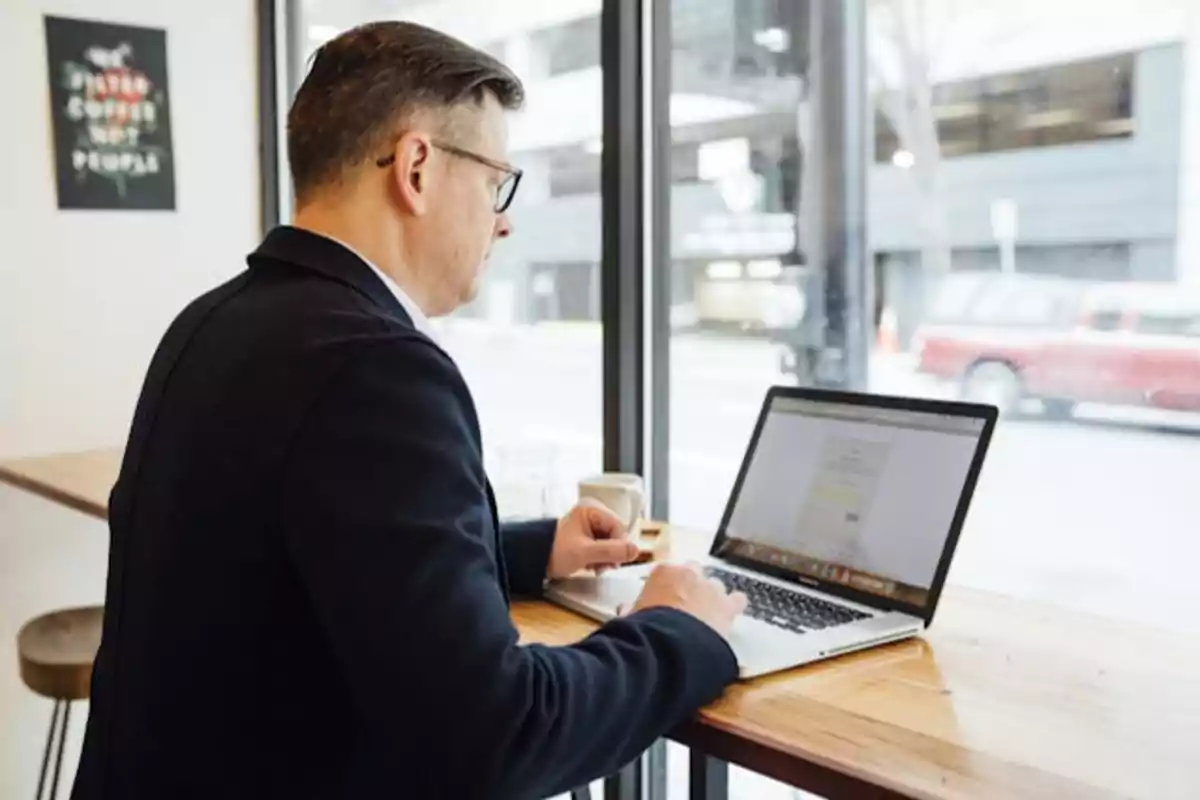 Hombre trabajando en una computadora portátil en una cafetería junto a una ventana.