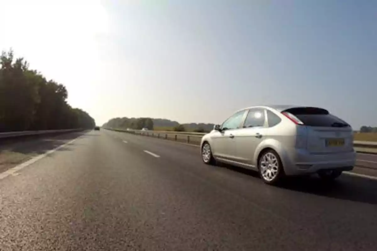 Un coche plateado conduciendo por una carretera despejada con árboles a un lado y cielo despejado.