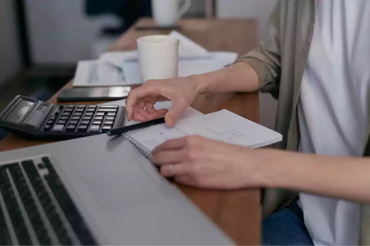 Persona trabajando en un escritorio con una computadora portátil, una calculadora y un cuaderno.