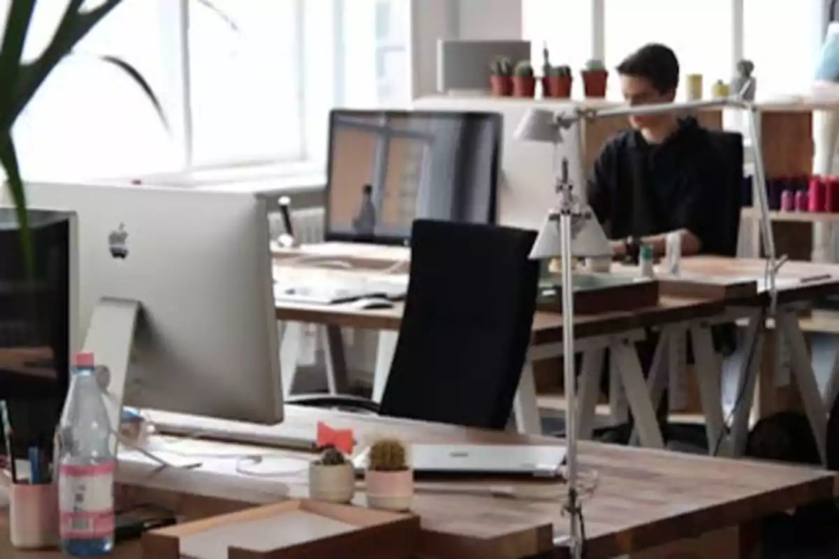 Oficina moderna con escritorios de madera, computadoras y plantas, con una persona trabajando al fondo.