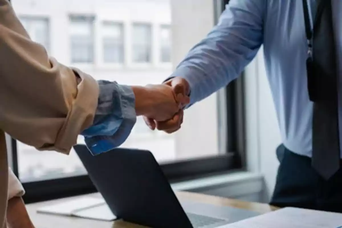 Dos personas dándose la mano en una oficina con una computadora portátil sobre la mesa.