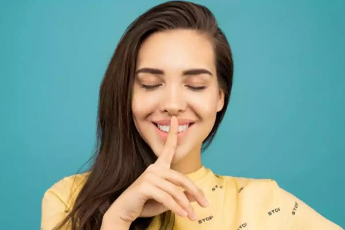 Mujer sonriendo con los ojos cerrados y haciendo el gesto de silencio con el dedo índice sobre los labios, fondo azul.